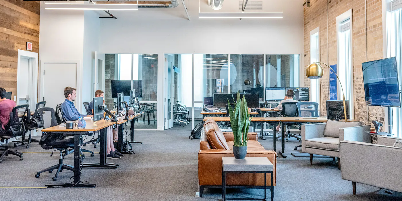 A bright open coworking office: people working at desks with laptops and monitors, exposed brick and timber, lounge seating and daylight